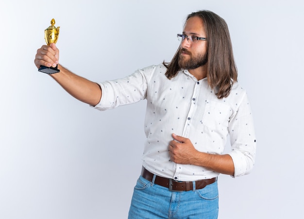 Bel homme adulte confiant portant des lunettes gardant la main sur le ventre qui s'étend et regarde la coupe du gagnant isolée sur le mur blanc