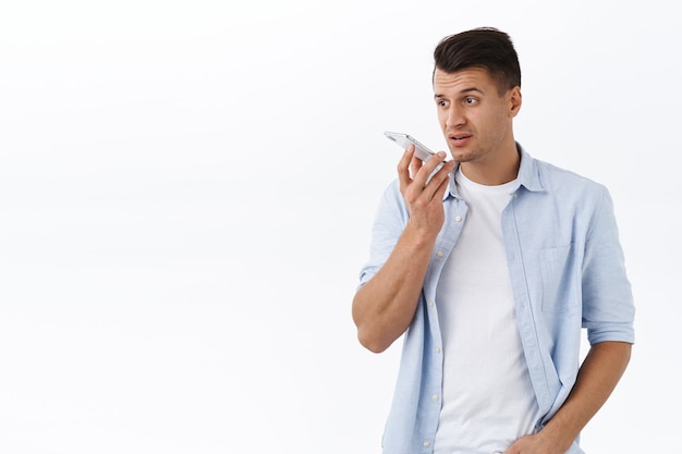 Un bel homme adulte occupé enregistre une messagerie vocale ou un message vocal sur un téléphone portable, tient le smartphone près des lèvres l'air sérieux et réfléchi, a une conversation, un mur blanc debout