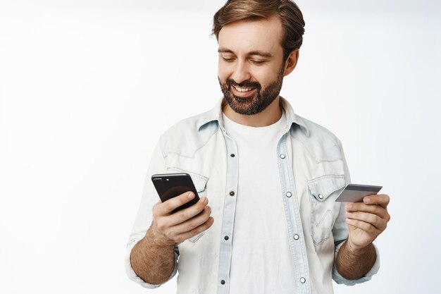 Bel homme adulte utilisant une carte de crédit et un téléphone portable regardant l'écran du smartphone avec un sourire heureux fond blanc