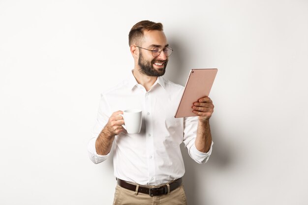 Bel homme d'affaires, boire du café et lire sur tablette numérique, souriant heureux, debout