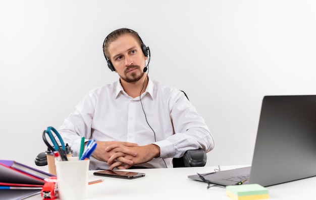 Photo gratuite bel homme d'affaires en chemise blanche et casque avec un microphone à côté avec un visage sérieux assis à la table en offise sur fond blanc