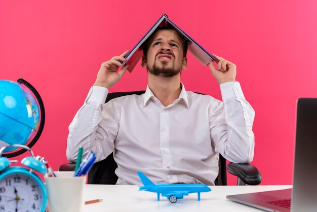 Bel homme d'affaires en chemise blanche avec globe holding notebook sur sa tête à la confusion assis à la table en offise sur fond rose