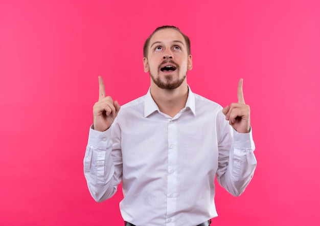 Bel homme d'affaires en chemise blanche regardant vers le haut pointant avec l'index debout heureux et positif sur fond rose