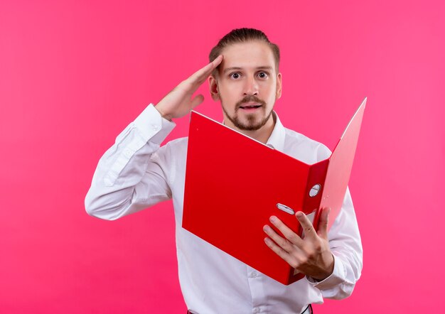 Bel homme d'affaires en chemise blanche tenant un dossier ouvert regardant la caméra surpris en saluant debout sur fond rose