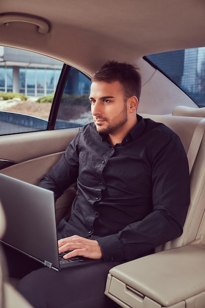 Un bel homme d'affaires en chemise noire travaillant avec un ordinateur portable à l'intérieur de la voiture.