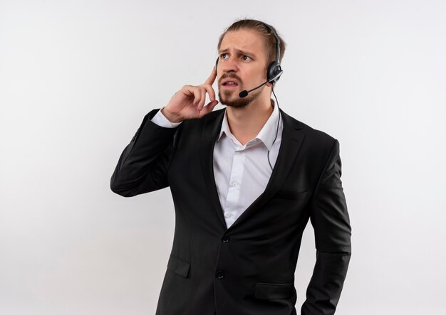 Bel homme d'affaires en costume et casque avec un microphone à l'écoute d'un client à la confusion debout sur fond blanc