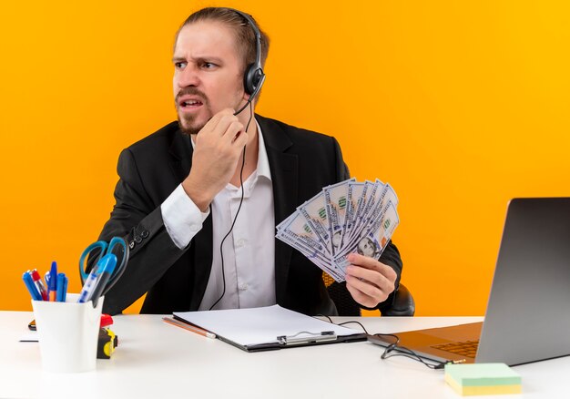 Bel homme d'affaires en costume et casque avec un microphone montrant cush à côté perplexe et mécontent assis à la table en offise sur fond orange