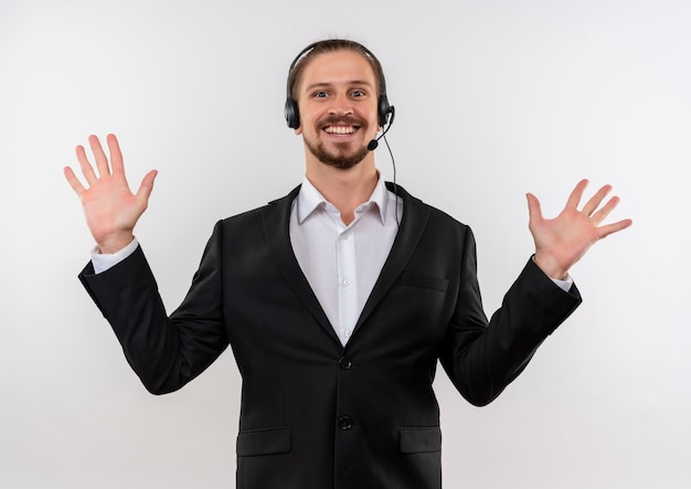 Bel homme d'affaires en costume et casque avec un microphone regardant la caméra en levant les paumes dans la reddition debout sur fond blanc