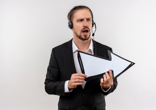 Photo gratuite bel homme d'affaires en costume et casque avec un microphone tenant une flèche blanche à côté surpris et confus debout sur fond blanc