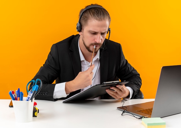 Bel homme d'affaires en costume et casque avec un microphone tenant le presse-papiers avec des documets à la recherche avec un visage sérieux assis à la table en offise sur fond orange