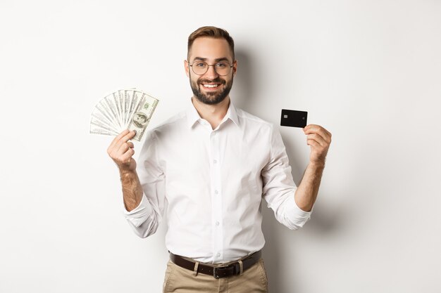 Bel homme d'affaires montrant la carte de crédit et l'argent en dollars, souriant satisfait, debout