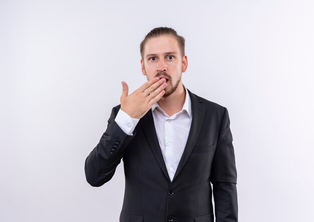 Bel homme d'affaires portant un costume à la bouche surprise et étonné couvrant avec la main debout sur fond blanc