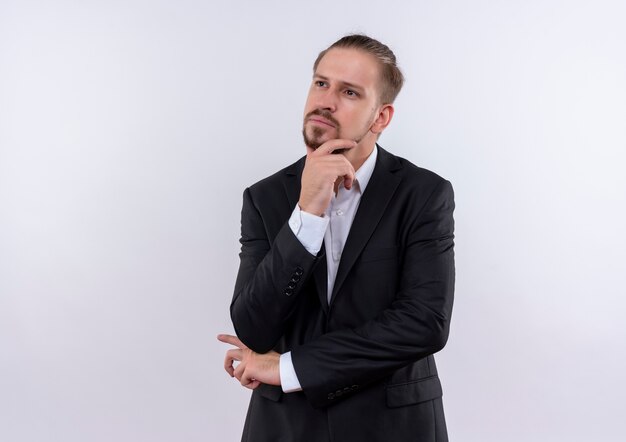 Bel homme d'affaires portant un costume à côté avec la main sur le menton avec une expression pensive debout sur fond blanc