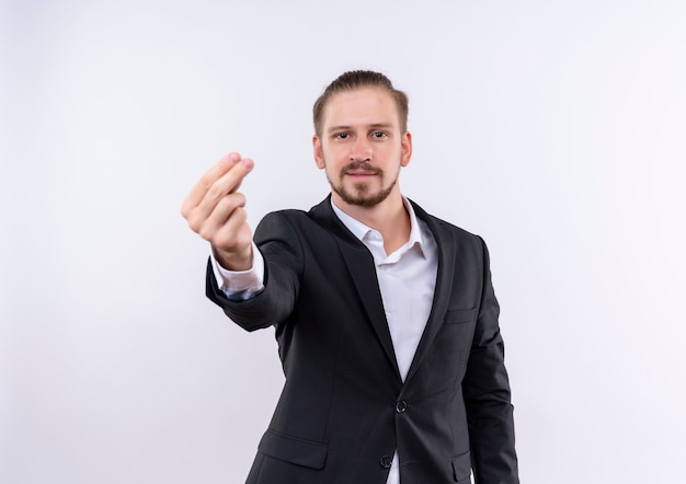 Bel homme d'affaires portant costume regardant la caméra en se frottant les doigts faisant le geste de l'argent debout sur fond blanc