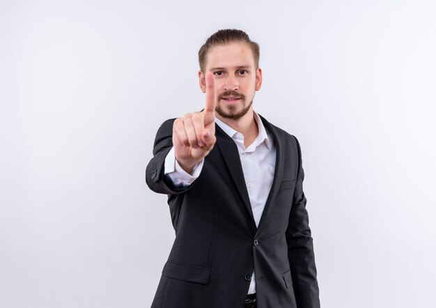 Bel homme d'affaires portant costume regardant la caméra en souriant montrant l'index debout sur fond blanc