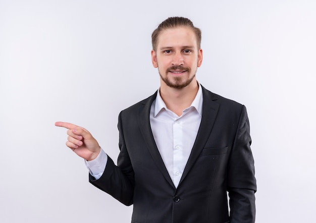 Bel homme d'affaires portant costume regardant la caméra avec un sourire confiant pointant avec l'index sur le côté debout sur fond blanc