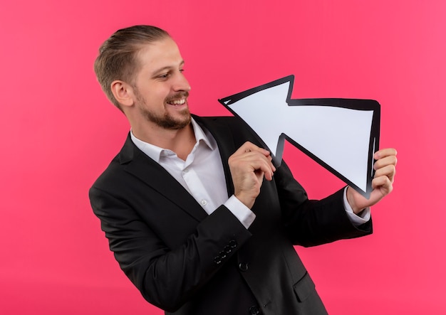 Bel homme d'affaires portant un costume tenant une flèche lokant à sourire debout sur fond rose