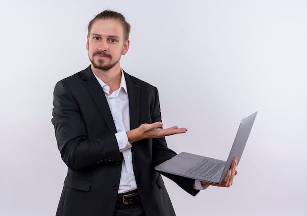Bel homme d'affaires portant costume tenant un ordinateur portable présentant le bras de la main à la confiance debout sur fond blanc