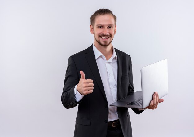 Bel homme d'affaires portant costume tenant des pages blanches regardant la caméra en souriant joyeusement montrant les pouces vers le haut debout sur fond blanc