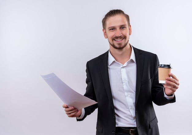 Bel homme d'affaires portant costume tenant une tasse de café et des pages blanches regardant la caméra en souriant joyeusement debout sur fond blanc