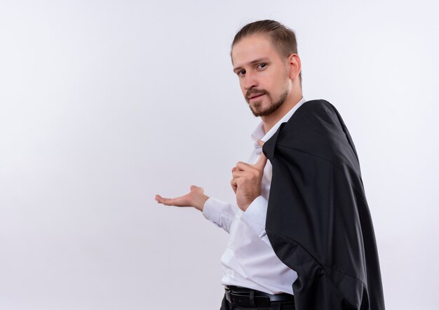 Bel homme d'affaires portant sa veste sur l'épaule souriant présentant quelque chose avec le bras de sa main debout sur fond blanc