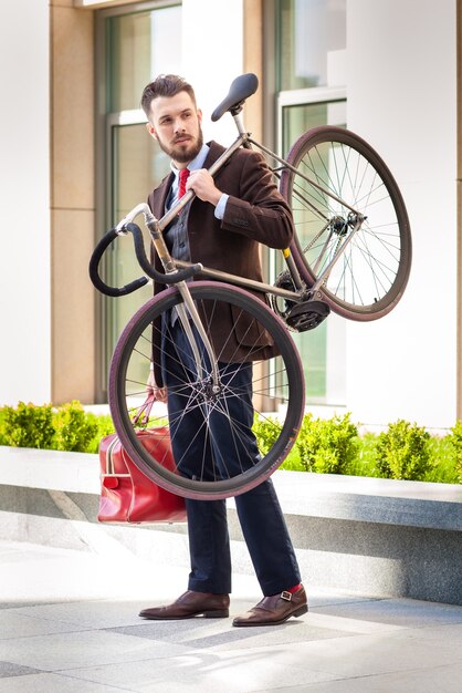 Bel homme d'affaires avec un sac rouge portant son vélo dans les rues de la ville.