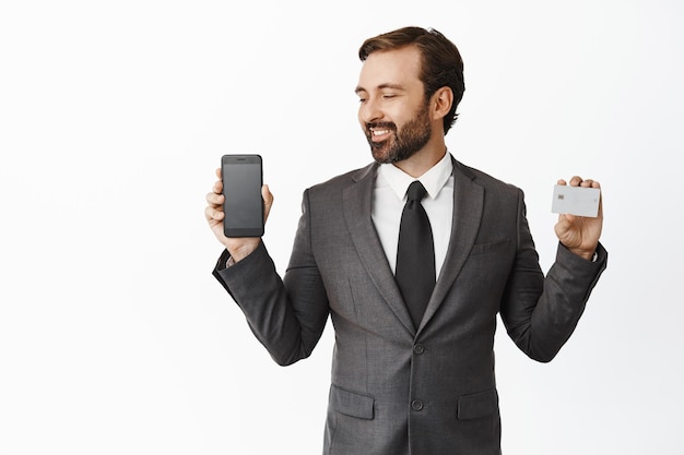 Bel homme d'affaires souriant regardant heureux l'écran du téléphone montrant l'interface de l'application smartphone et le fond blanc de la carte de crédit