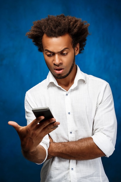 Bel homme africain mécontent regardant téléphone sur mur bleu.