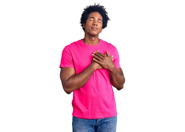 Bel homme afro-américain aux cheveux afro portant un t-shirt rose décontracté souriant avec les mains sur la poitrine avec les yeux fermés et un geste reconnaissant sur le concept de santé du visage
