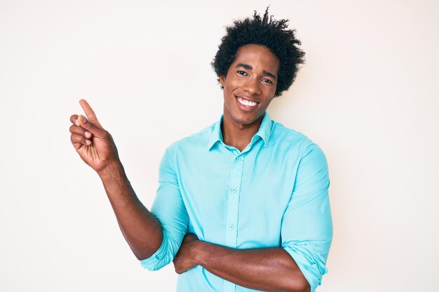 Bel homme afro-américain aux cheveux afro portant des vêtements décontractés avec un grand sourire sur le visage pointant avec la main et le doigt sur le côté en regardant la caméra