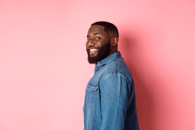 Bel homme afro-américain avec barbe, tourner le visage devant la caméra et souriant confiant, debout sur fond rose