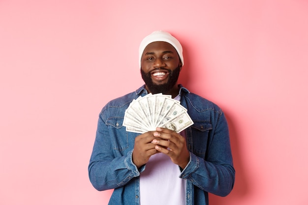 Bel homme afro-américain barbu montrant de l'argent, gagnant des dollars, debout sur fond rose