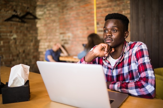 Bel homme afro-américain dans des vêtements décontractés tenant une tasse de café et utilisant un ordinateur portable.