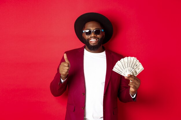 Bel homme afro-américain moderne dans des lunettes de soleil et des vêtements de fête, montrant le pouce avec des dollars, gagne de l'argent et a l'air satisfait, fond rouge.