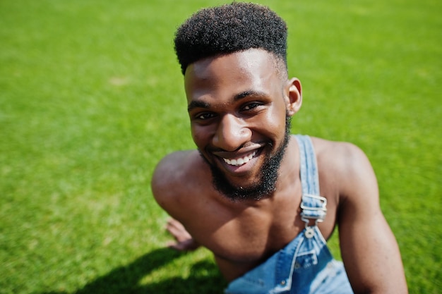 Bel Homme Afro-américain Sexy Torse Nu à La Salopette De Jeans Assis à L'herbe Verte Du Terrain De Football Du Stade Portrait D'homme Noir à La Mode