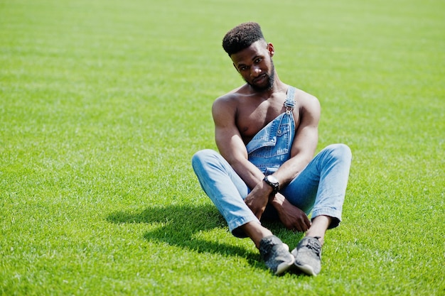 Photo gratuite bel homme afro-américain sexy torse nu à la salopette de jeans assis à l'herbe verte du terrain de football du stade portrait d'homme noir à la mode