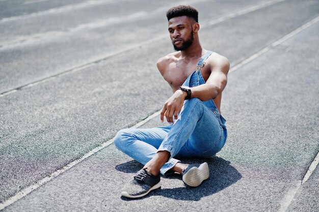 Bel homme afro-américain sexy torse nu à la salopette de jeans assis à l'hippodrome du stade Portrait d'homme noir à la mode