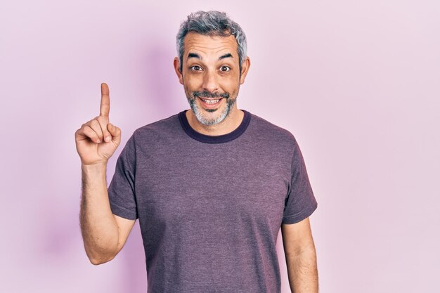 Bel homme d'âge moyen aux cheveux gris portant un t-shirt décontracté pointant le doigt vers le haut avec une idée réussie. excité et heureux. numéro un.