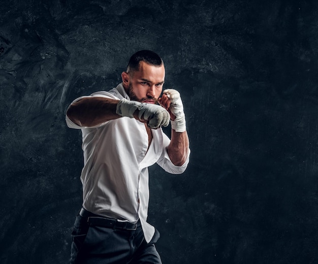 Photo gratuite un bel homme agressif en chemise blanche fait la démonstration de son coup de poing dans un studio photo sombre.