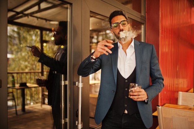 Bel homme arabe bien habillé fume un cigare avec un verre de whisky au balcon du pub