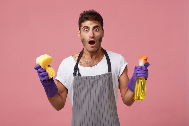 Bel homme ayant le visage sale portant un tablier et des gants tenant une éponge et un spray de nettoyage ayant une expression choquée réalisant combien il doit nettoyer. Homme perplexe faisant les tâches ménagères isolées