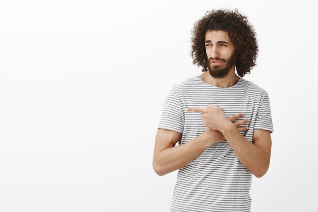 Bel homme barbu adulte avec une coiffure afro, pointant et regardant à droite avec dégoût et doute, mec moqueur sans croire en sa victoire