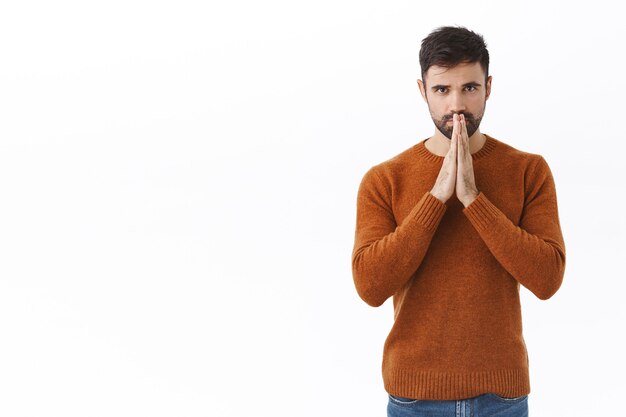 Bel homme barbu à l'air sérieux, inquiet et concerné, priant pour la guérison mondiale de covid19, se tenant la main près des lèvres en attendant des nouvelles ou des résultats importants, mur blanc