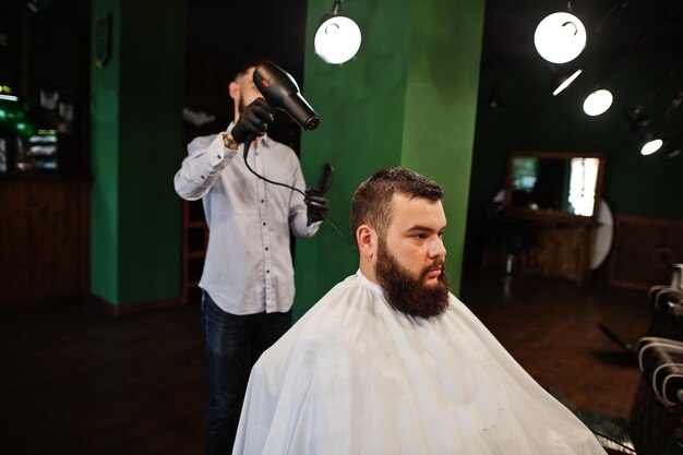 Bel homme barbu au salon de coiffure coiffeur au travail à l'aide d'un sèche-cheveux