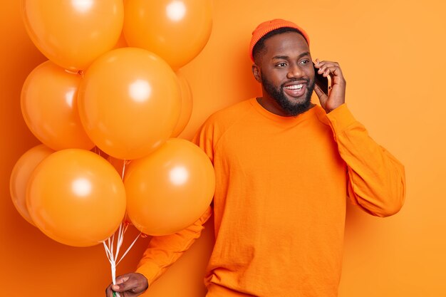 Bel homme barbu a une conversation heureuse via smartphone célèbre un événement positif invite des amis à la fête décore la chambre avec des ballons isolés sur un mur orange