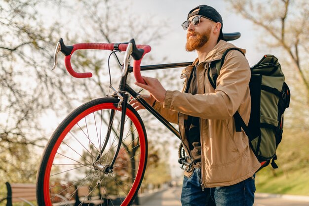 Bel homme barbu élégant hipster en veste et lunettes de soleil marchant seul dans la rue avec sac à dos sur vélo routard voyageur de mode de vie actif sain