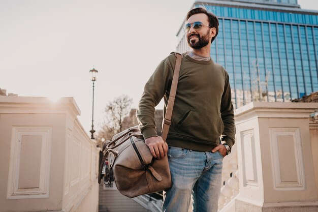 Bel homme barbu élégant marchant dans la rue de la ville avec sac de voyage en cuir portant un sweat-shirt et des lunettes de soleil, tendance de style urbain, journée ensoleillée, confiant et souriant