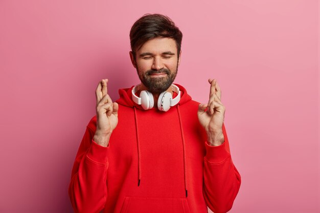 Un bel homme barbu persistant se tient avec les doigts croisés, ferme les yeux et attend un moment spécial, porte un sweat-shirt rouge et des écouteurs stéréo autour du cou, attend les résultats, pose seul à l'intérieur