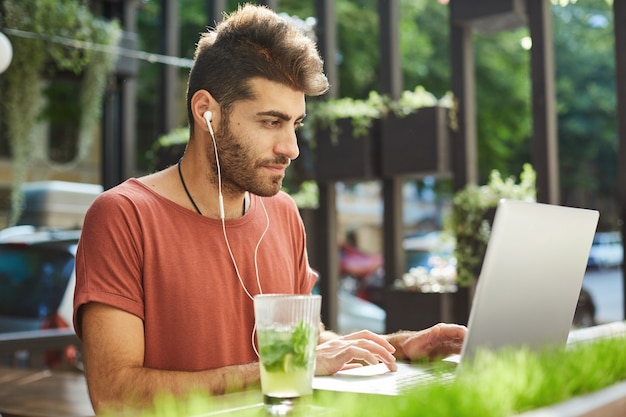 Bel homme barbu, pigiste travaillant à distance du café en plein air, programmeur avec ordinateur portable écoutant de la musique pour se concentrer sur le travail