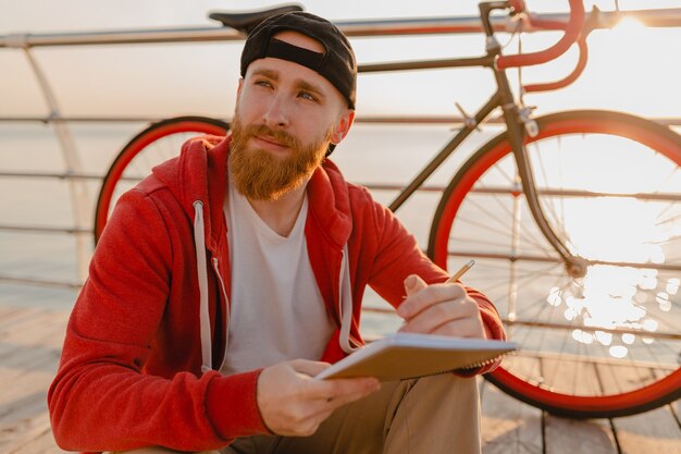 Bel homme barbu de style hipster pensant en sweat à capuche rouge étudiant en ligne pigiste écrit en prenant des notes avec vélo au lever du soleil du matin au bord de la mer